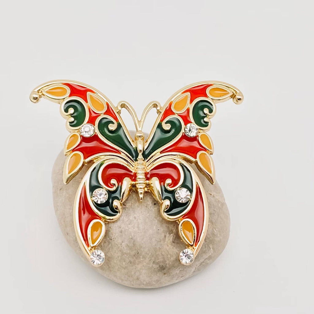 Butterfly-shaped brooch with red, green, and gold colors on a light gray background