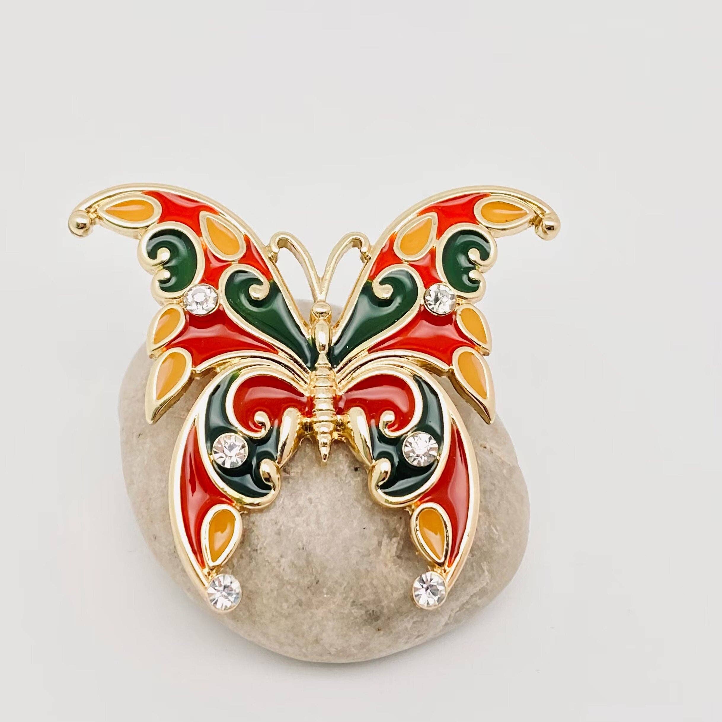 Butterfly-shaped brooch with red, green, and gold colors on a light gray background