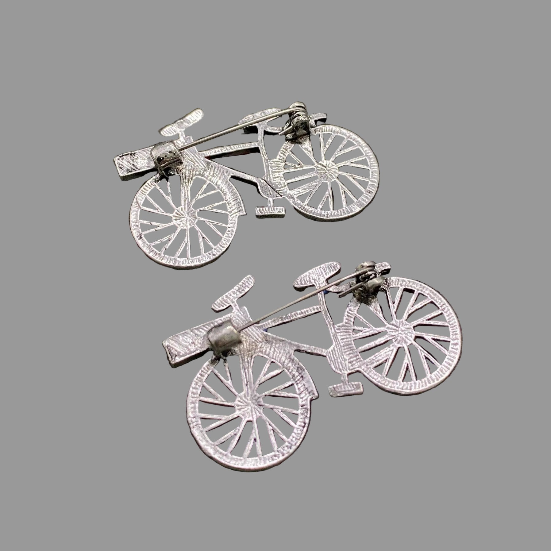 Back view of two bicycle shaped brooches, showing the clasp mechanism