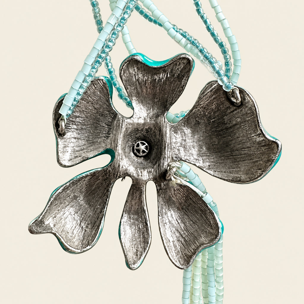 A backside view of a flower-shaped pendant necklace with a textured silver-toned metal flower and light blue beaded strands on a beige fabric background.