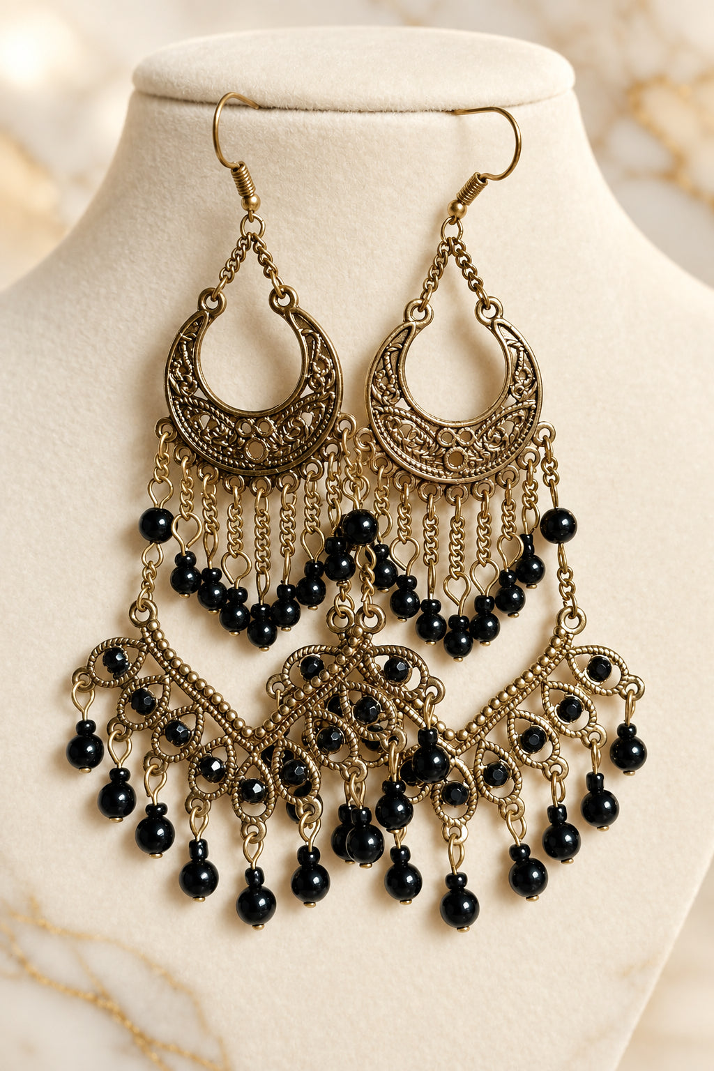 A pair of ornate dangling earrings displayed on a cream jewelry bust. The earrings feature gold-toned filigree crescent moon and floral motifs with multiple tiers of small black beads.