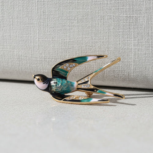 A swallow-shaped brooch with black, white, and blue enamel details and clear rhinestone accents on the wings, shown on a beige background