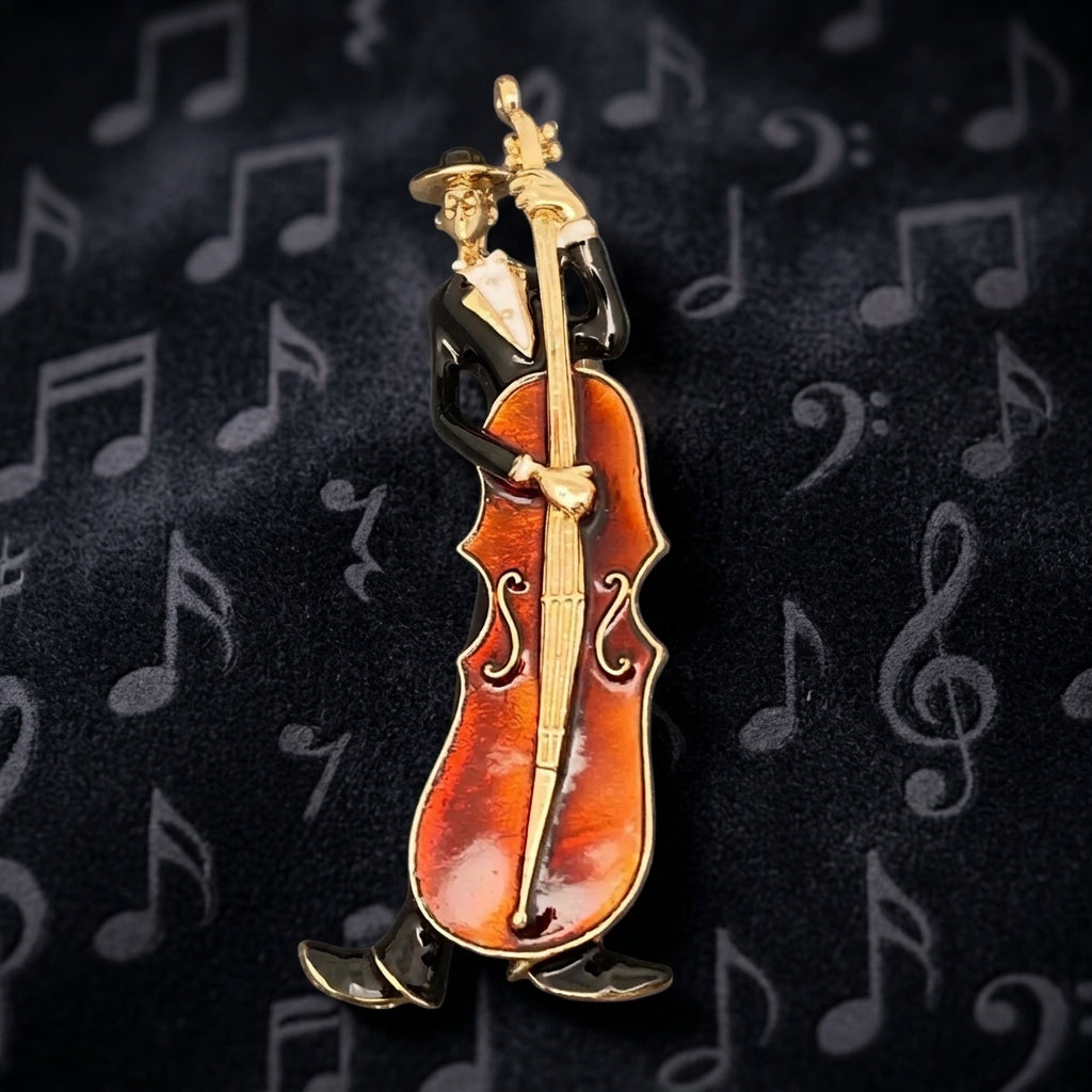 A cellist brooch featuring a gold-tone figure in a black suit, playing a brown enamel cello, set against a dark background with patterned musical notes.