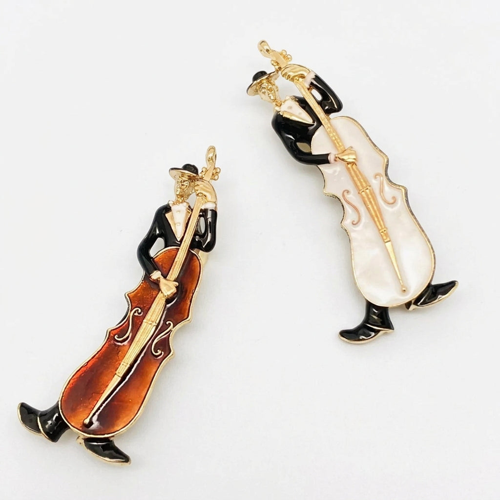 Two cellist-shaped brooches on a plain white background. One brooch features a brown enamel cello with gold-tone metal, and the other features a white enamel cello with gold-tone metal.