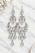 A pair of silver-toned chandelier drop earrings with multiple clear faceted rhinestones, displayed against a white marble background.