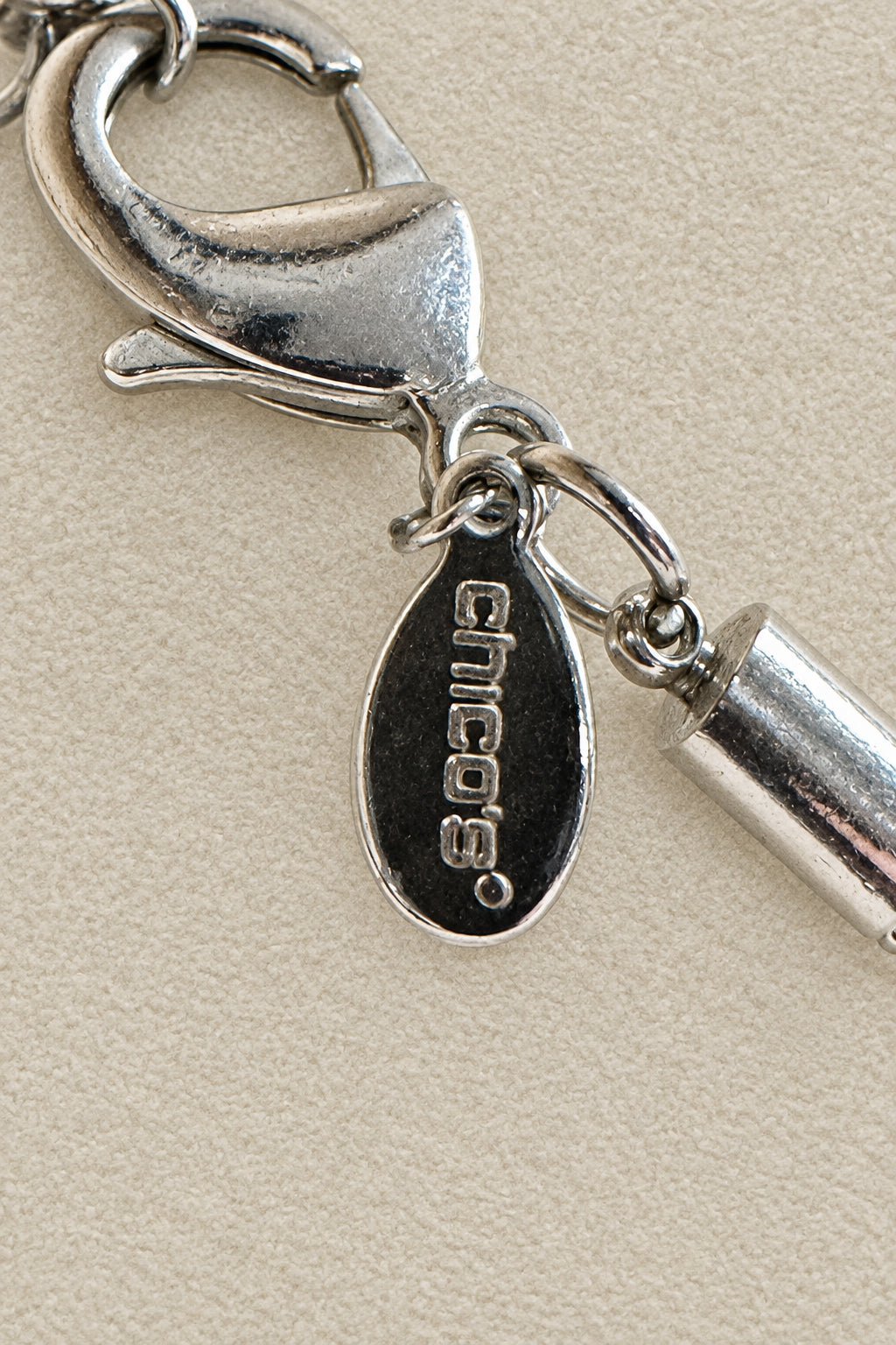A close-up of a silver-toned lobster clasp and Chico's designer jewelry tag, set against a textured beige background.