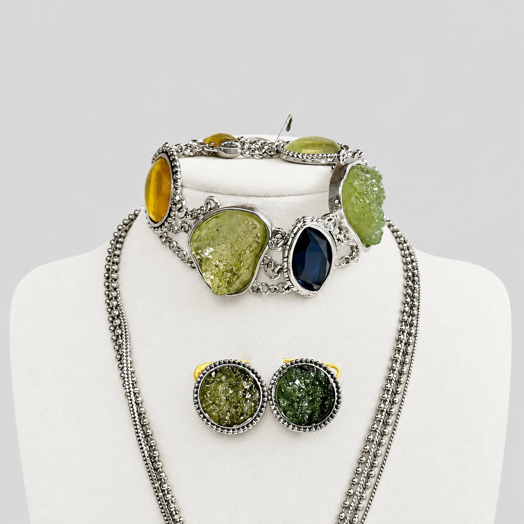 A jewelry set displayed on a cream-colored mannequin bust, featuring a silver-tone chain necklace, a matching bracelet, and a pair of stud earrings with textured green and yellow stones in silver settings.