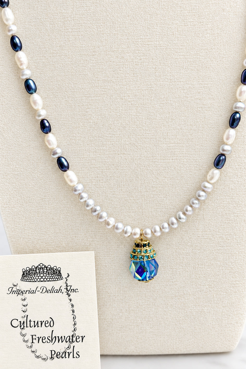 A freshwater pearl necklace with dark blue oval pearls and a blue faceted crystal pendant, displayed on a beige jewelry bust with a brand card for 'Folklorial Delights Inc.'.