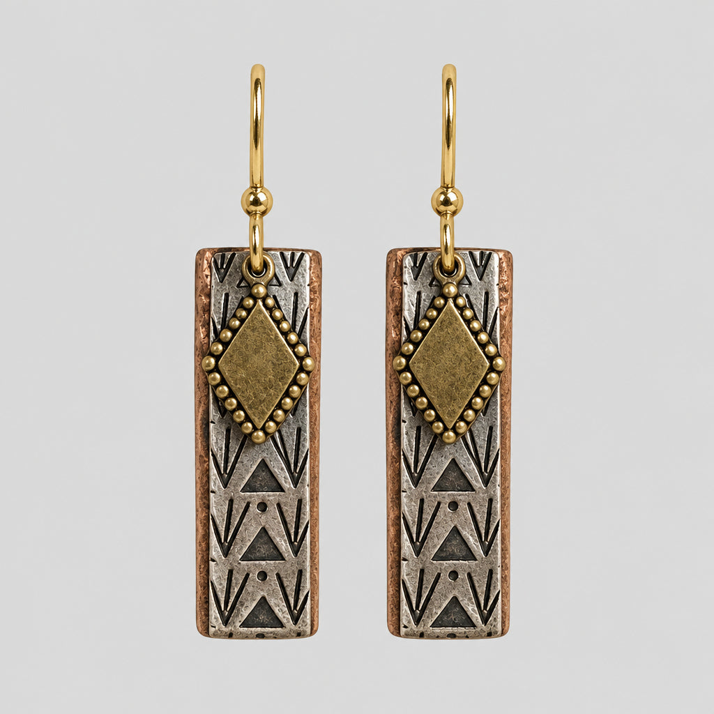A pair of mix metal rectangular drop earrings with textured silver-toned geometric patterns, a gold-toned diamond accent, and gold-toned fishhook wires, displayed on a textured beige surface.