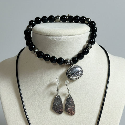 A jewelry set displayed on a cream fabric bust, featuring a black cord necklace, a black beaded bracelet with a silver charm, and a matching pair of silver textured dangling earrings.