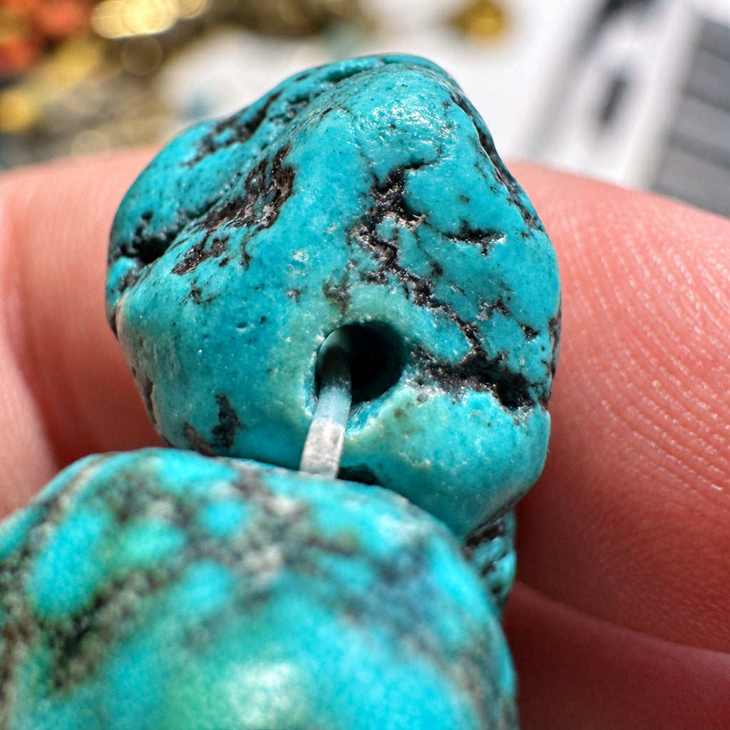 A close-up of a turquoise-colored stone bead or jewelry component with a hole, held between fingers against a blurred background.