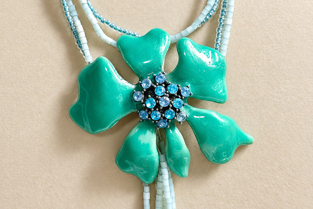 A turquoise enamel flower pendant necklace with a central cluster of blue rhinestones, suspended from white and light blue beaded cords on a neutral beige background.