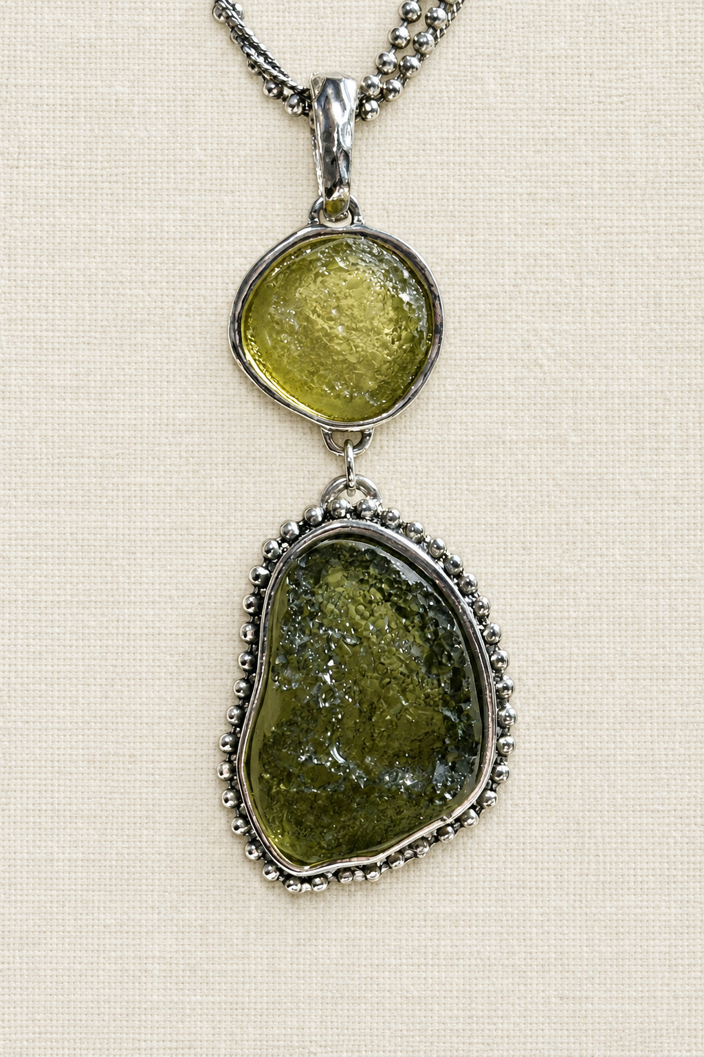 A silver pendant necklace featuring two stacked oval gemstone sections, one with a yellow-green stone and the other with a larger olive-green stone, displayed on a textured beige background.