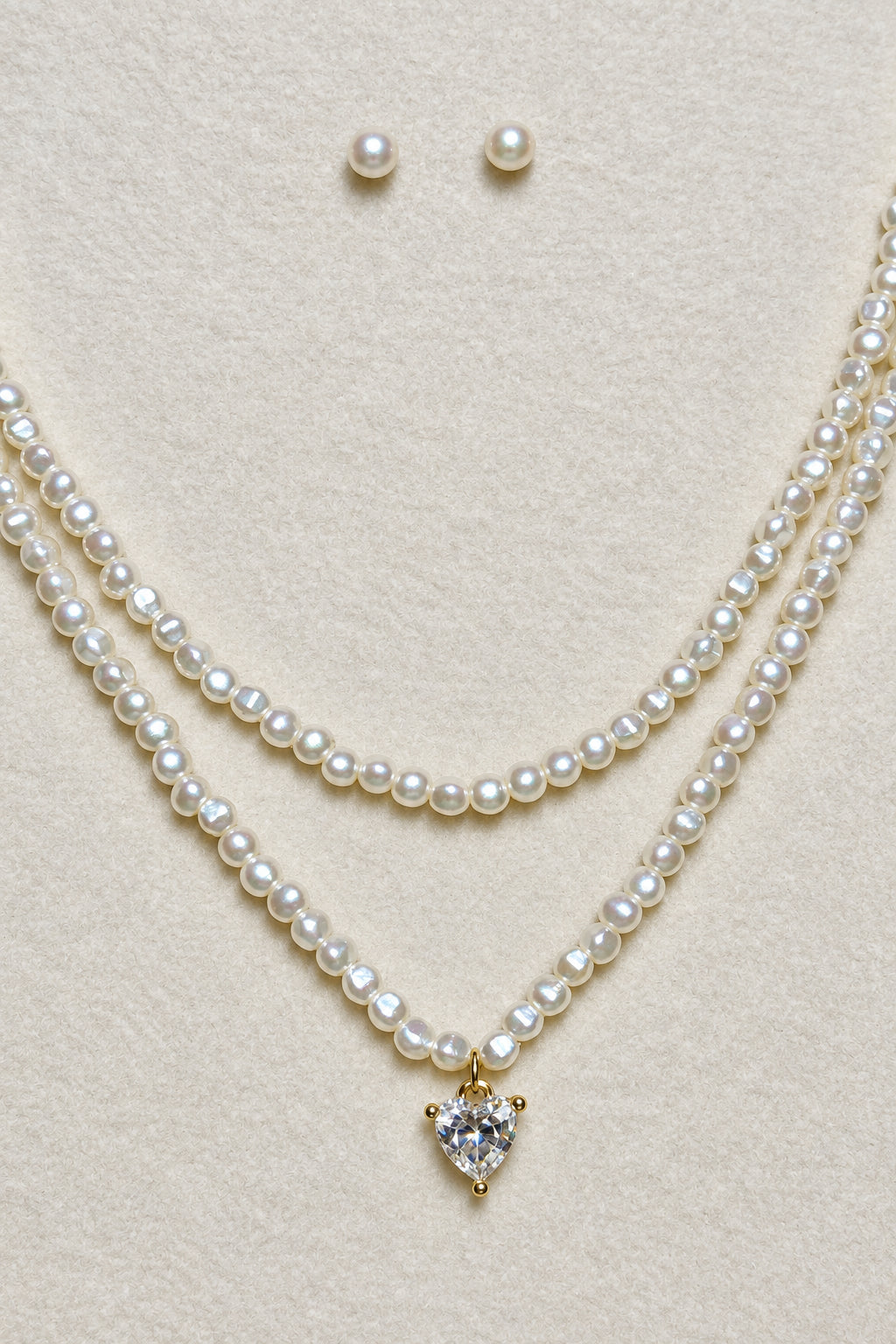 A double-strand pearl necklace with a small heart-shaped crystal pendant, displayed on a textured beige jewelry bust alongside matching pearl stud earrings.