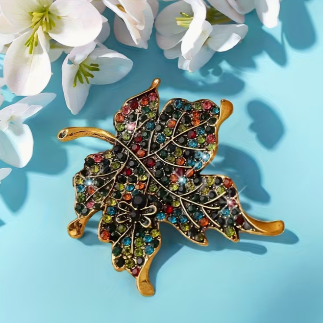 Leaf-shaped brooch with colorful gemstones on a blue background with white flowers.