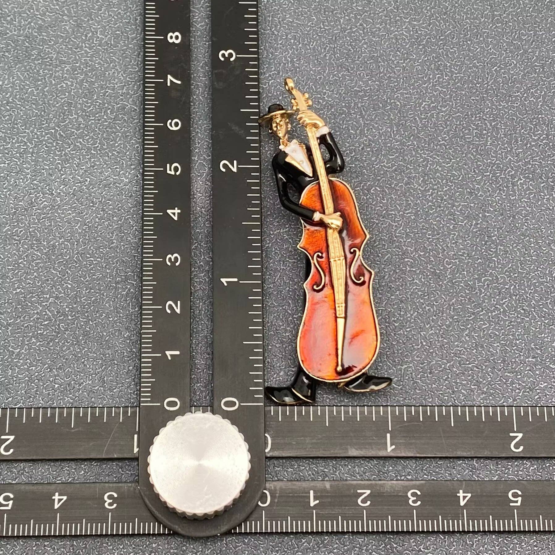 Decorative pin of a musician playing a cello, measured against a ruler on a gray background