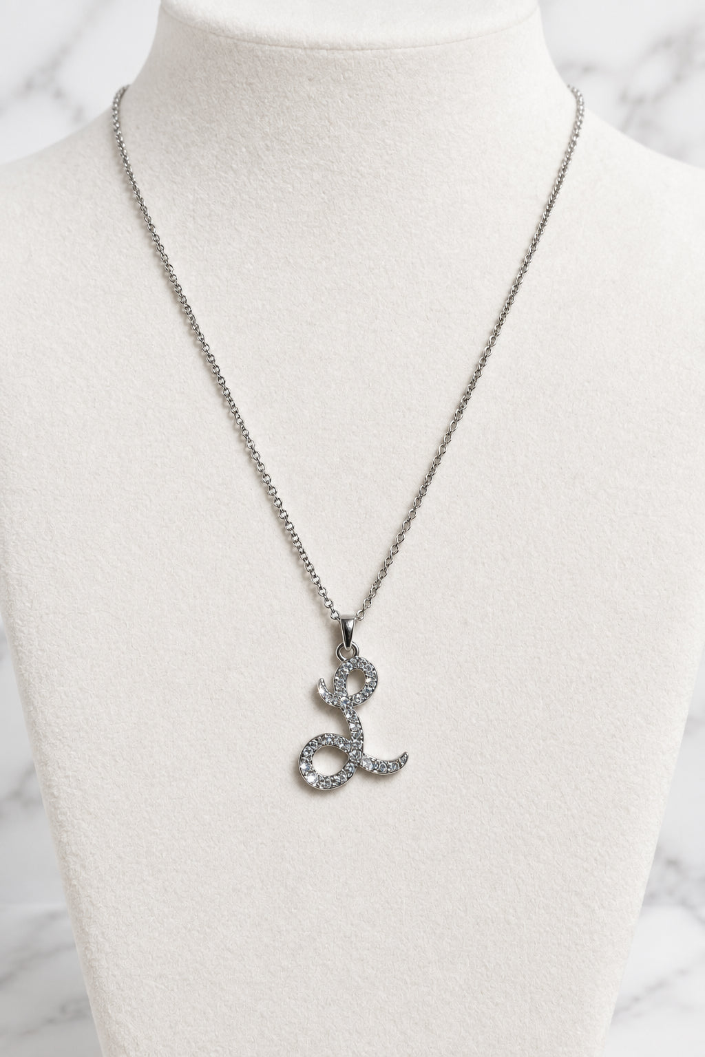 A silver-tone chain necklace with a stylized letter 'L' pendant encrusted with small clear stones, displayed on a cream-colored jewelry bust.