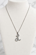 A silver-tone chain necklace with a stylized letter 'L' pendant encrusted with small clear stones, displayed on a cream-colored jewelry bust.
