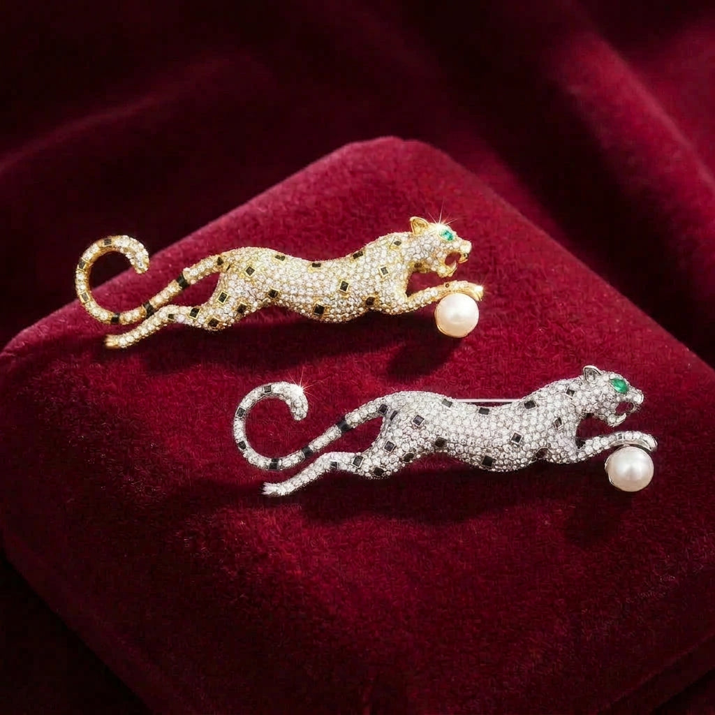 Two cheetah brooches displayed on a burgundy velvet surface. One brooch is gold and one is silver, both with pavé-set clear rhinestones, black spot accents, green crystal eyes, and a white pearl at its paw.