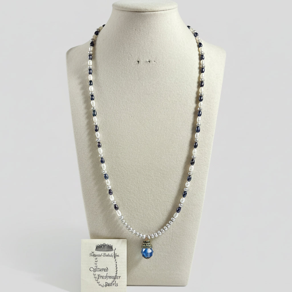 A freshwater pearl beaded necklace displayed on a cream fabric mannequin bust. The necklace features alternating strands of white pearls and dark navy blue pearls, with a central blue faceted crystal pendant.