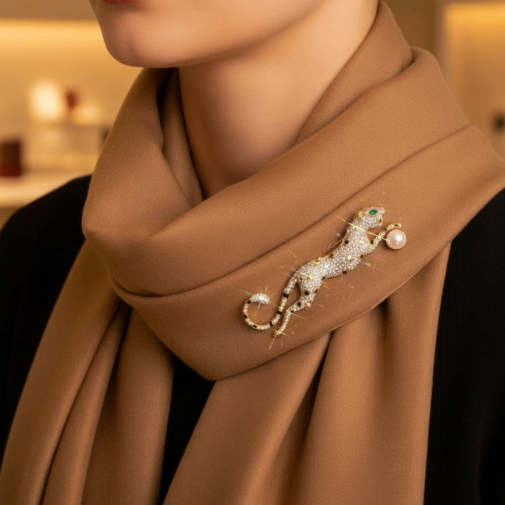 A close-up of a woman wearing a warm brown scarf with a jeweled cheetah brooch pinned near the collar. The brooch is embellished with rhinestones and a pearl, and the background is softly blurred.