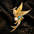 Jeweled phoenix bird brooch with gold-tone wings, clear rhinestones, blue floral details, and faux pearl, on a brown glittery  surface.