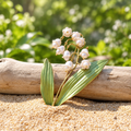 A lily of the valley floral brooch with white bell-shaped blossoms and green leaves, placed against a piece of driftwood in a blurred garden background.