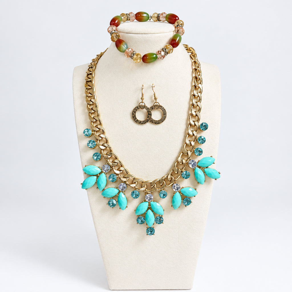 A gold-tone jewelry set displayed on a cream mannequin bust, featuring a statement necklace with turquoise and clear rhinestones, matching drop earrings, and a beaded bracelet with red, green, and gold beads, set against a light gray background.