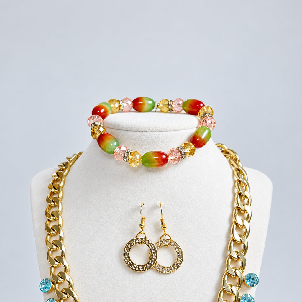 A jewelry set displayed on a cream mannequin bust, featuring a beaded bracelet with red, green, and gold beads, a pair of gold-tone textured hoop earrings, and layered gold chain necklaces, set against a light gray  background.