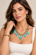 A woman wearing a gold-tone statement necklace featuring turquoise and clear rhinestones, matching gold drop earrings, and a beaded bracelet with red, green, and gold beads.