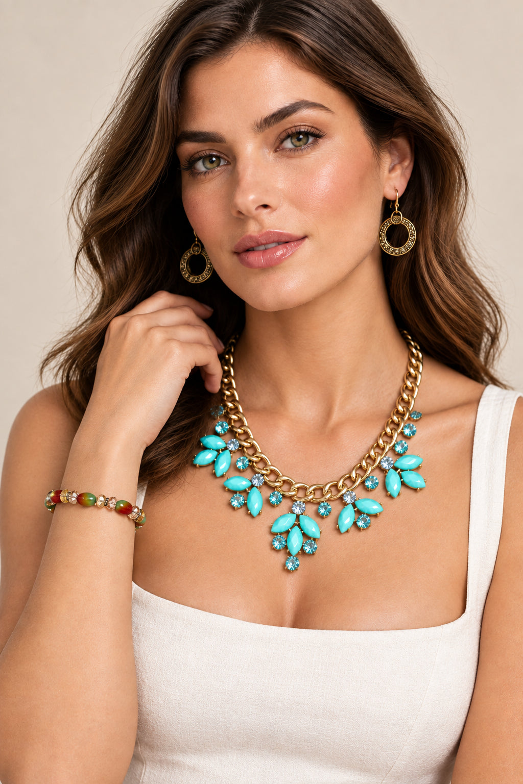 A woman wearing a gold-tone statement necklace featuring turquoise and clear rhinestones, matching gold drop earrings, and a beaded bracelet with red, green, and gold beads.