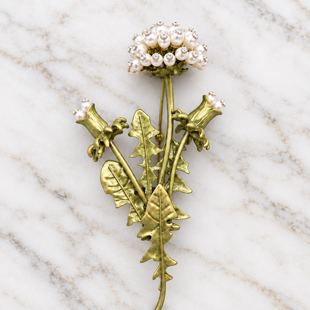 Dandelion brooch with white pearl-like blossoms and iridescent rhinestones, attached to metallic gold-green stems and leaves, against a marble background.