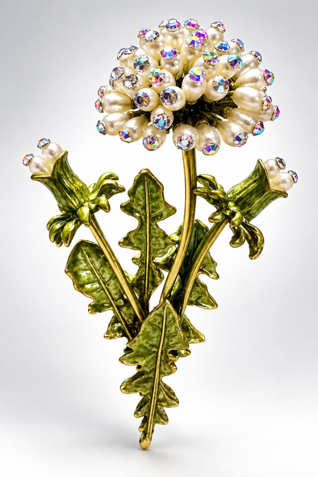 A close-up of a dandelion brooch with white pearl-like blossoms and iridescent rhinestones, attached to metallic gold-green stems and leaves, against a beige background.