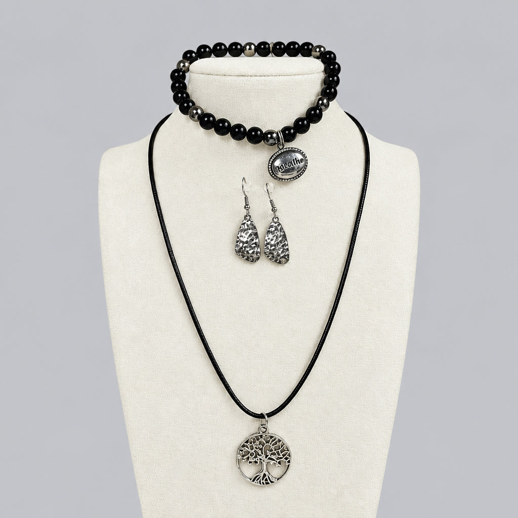 A jewelry set displayed on a cream fabric bust, featuring a black cord necklace with a matching silver tree pendant, a black beaded bracelet with a silver charm, and a pair of textured silver teardrop earrings, set against a light gray background.