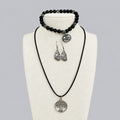 A jewelry set displayed on a cream fabric bust, featuring a black cord necklace with a matching silver tree pendant, a black beaded bracelet with a silver charm, and a pair of textured silver teardrop earrings, set against a light gray background.