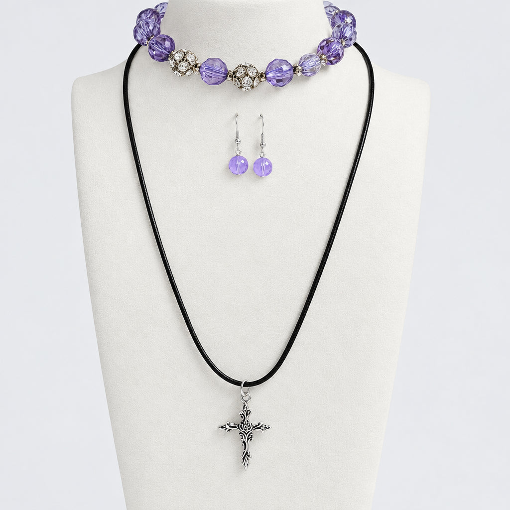 A jewelry set displayed on a cream fabric bust, featuring a black cord necklace with a silver-toned cross pendant, a matching purple beaded bracelet and a pair of dangling purple beaded earrings. Set against a light gray background.