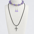 A jewelry set displayed on a cream fabric bust, featuring a black cord necklace with a silver-toned cross pendant, a matching purple beaded bracelet and a pair of dangling purple beaded earrings. Set against a light gray background.