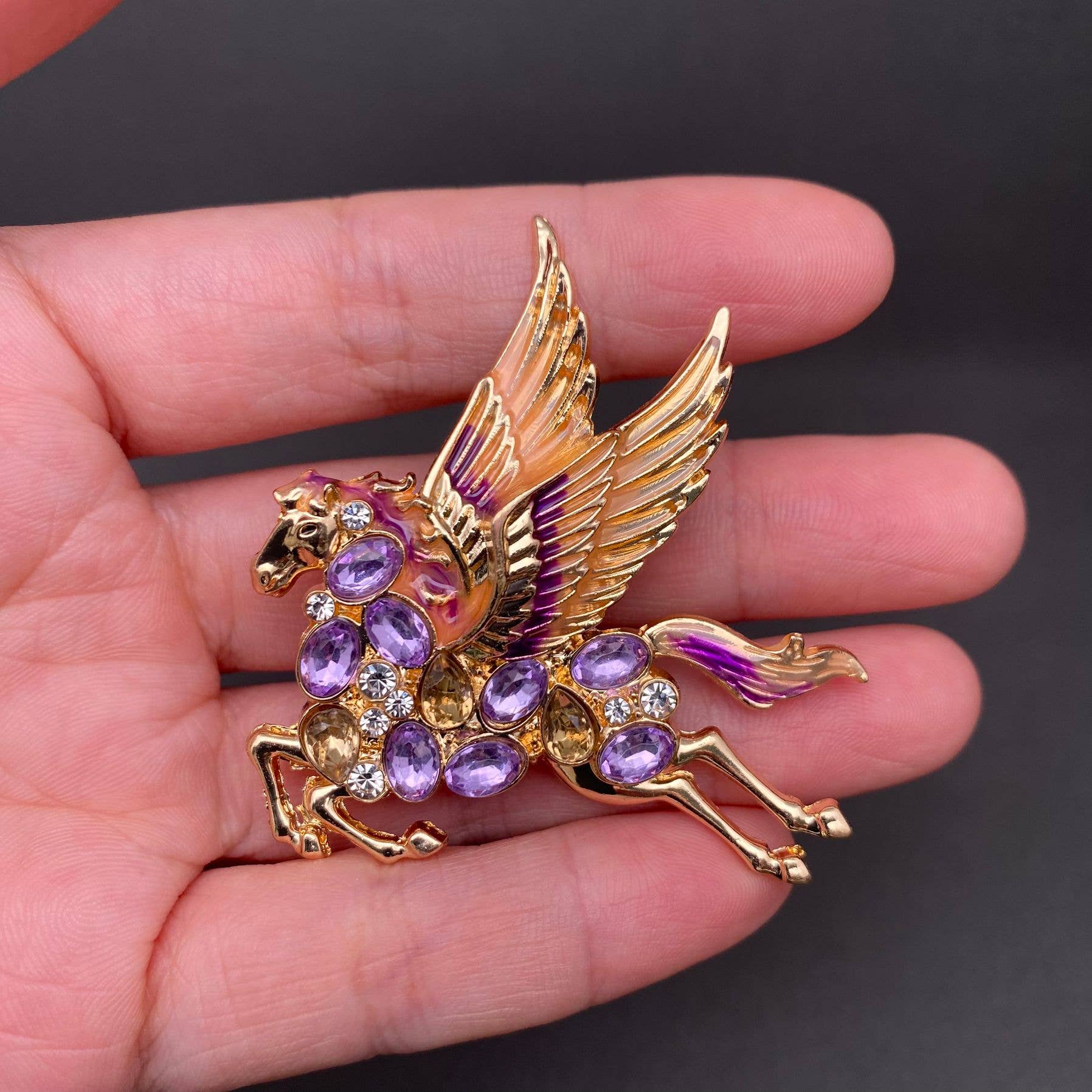Gold pegasus brooch with purple and violet gemstones held in a hand against a dark background