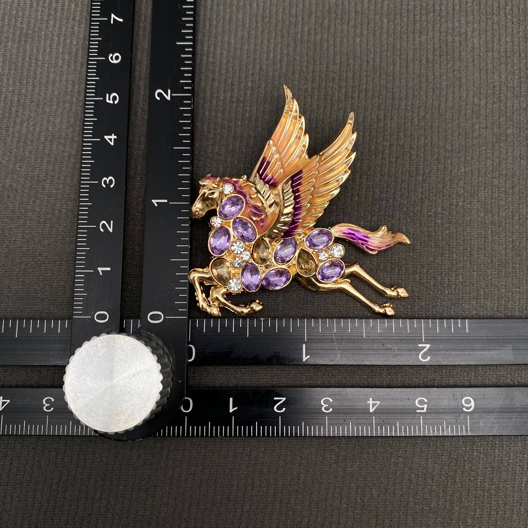 Gold pegasus brooch with purple and violet gemstones, measured against a ruler on a dark background