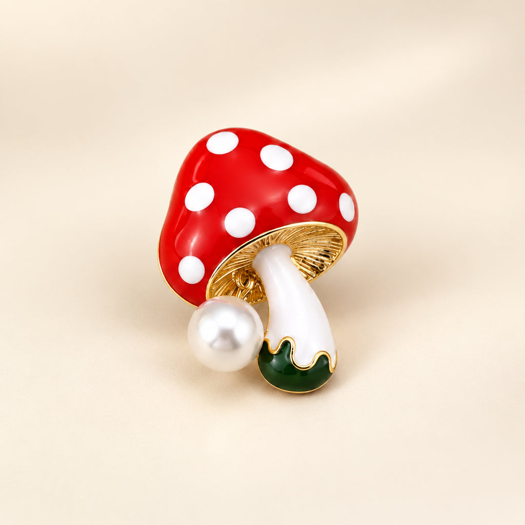 A red enamel mushroom brooch with white spots and a pearl accent, against a white background.