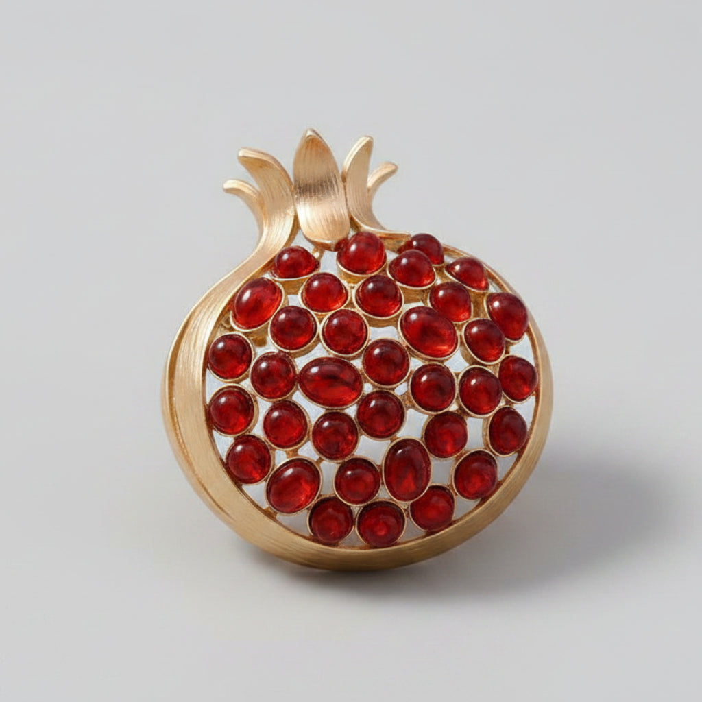 A gold-toned brooch shaped like a pomegranate, with a round cluster of red gemstones, displayed on gray background