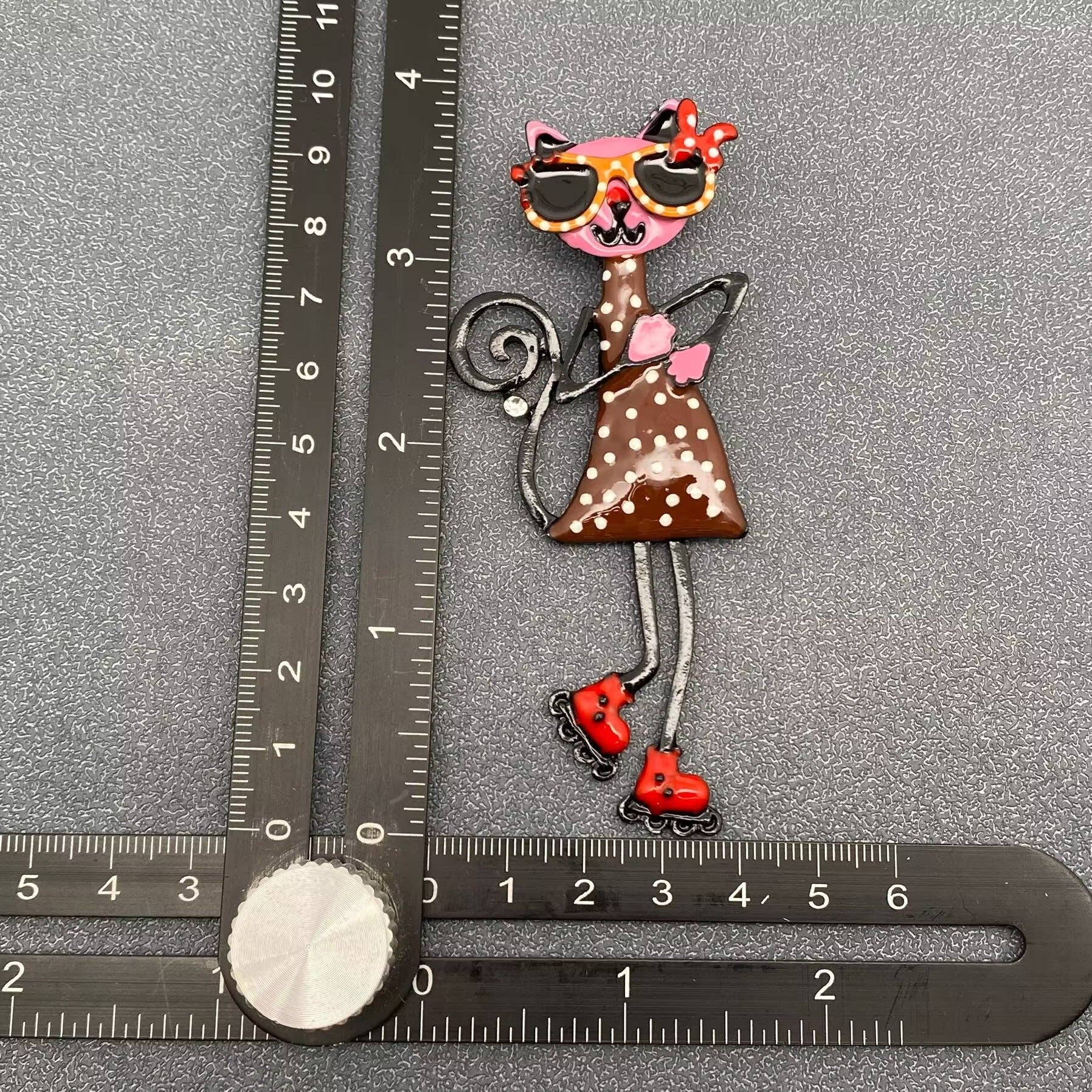 Decorative brooch of a polka dot cat with sunglasses and roller skates, measured against a ruler on a gray background