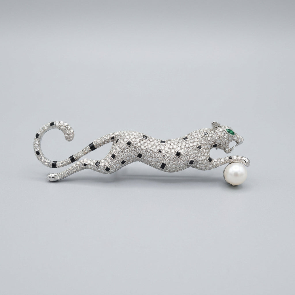 A silver cheetah brooch featuring pavé-set rhinestones, black stone spots, a green crystal eye, and a white pearl at its paw. Shown against a neutral gray background.
