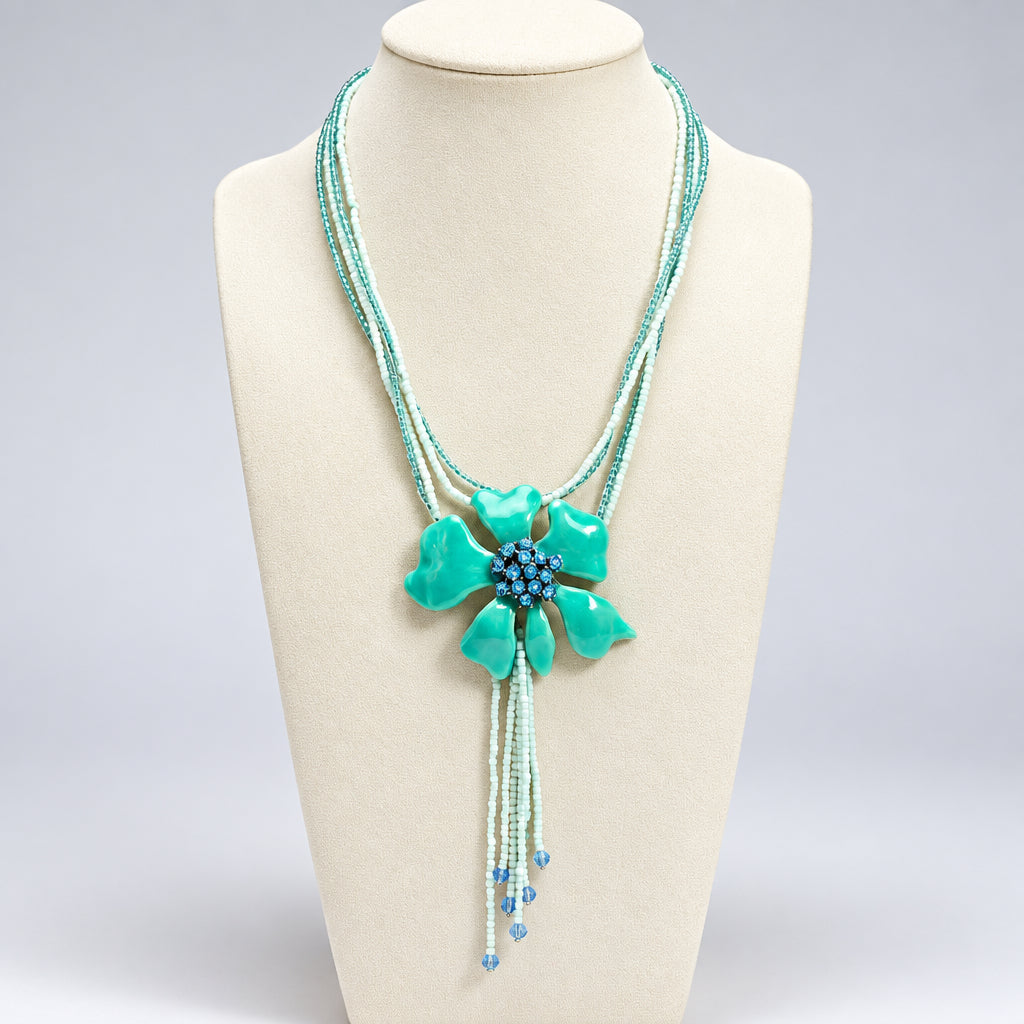 A turquoise and light blue beaded necklace displayed on a cream fabric mannequin bust. The necklace features a large, textured turquoise enamel flower centerpiece with a blue rhinestone-embellished center, surrounded by multiple strands of light blue and white beads.