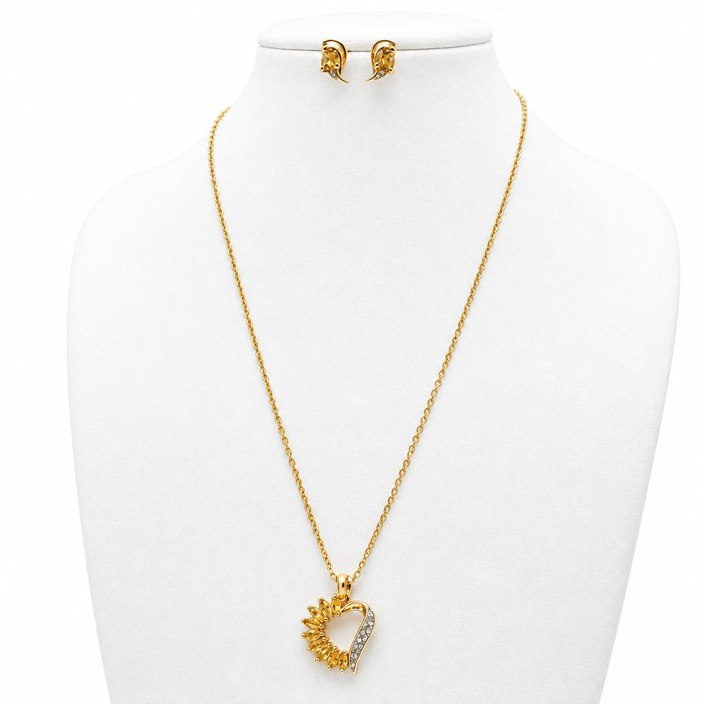 Gold necklace and matching stud earrings displayed on a cream jewelry bust. The necklace features a heart-shaped pendant with a sunburst design and a small rhinestone inlay.