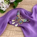 Colorful butterfly-shaped brooch with gemstones, on a purple satin scarf with flowers in the background