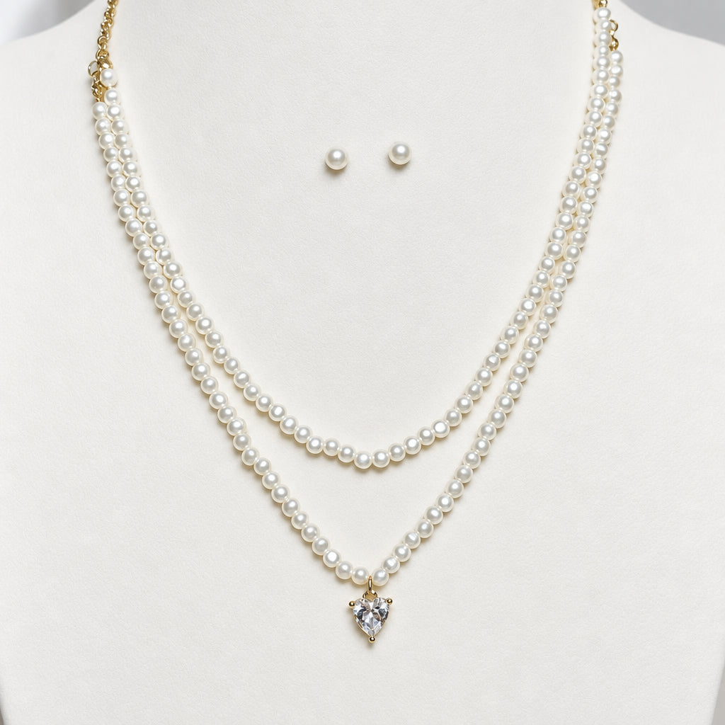 A double-strand pearl necklace with a small heart-shaped crystal pendant, displayed on a cream-colored jewelry bust alongside matching pearl stud earrings.