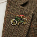 Bicycle-shaped brooch with gold frame, green body, and red seat pinned on a brown jacket